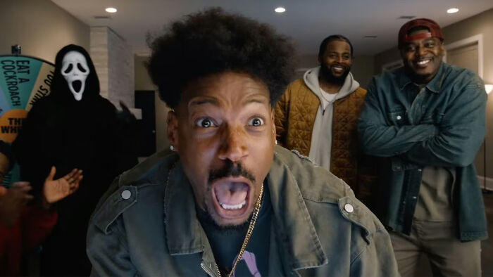 Um homem grita em primeiro plano, com Ghostface e dois homens sorridentes ao fundo. UM 