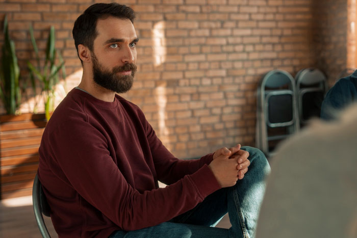 Bearded man in a maroon shirt sitting thoughtfully indoors, representing SIL feeding info to unstable ex in a tense setting.