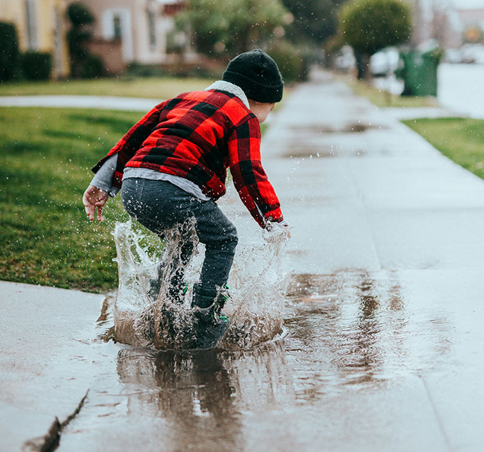 Child in school clothes jumping and splashing water in a puddle outdoors, capturing a moment of playful weirdo behavior.