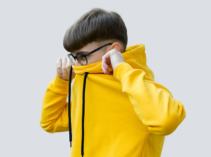 Teen kid in yellow hoodie covering face, wearing glasses, representing school kids who earned the weirdo title.
