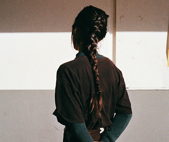 A school kid with a long braid stands facing a wall, representing stories of kids labeled as weirdo in school.