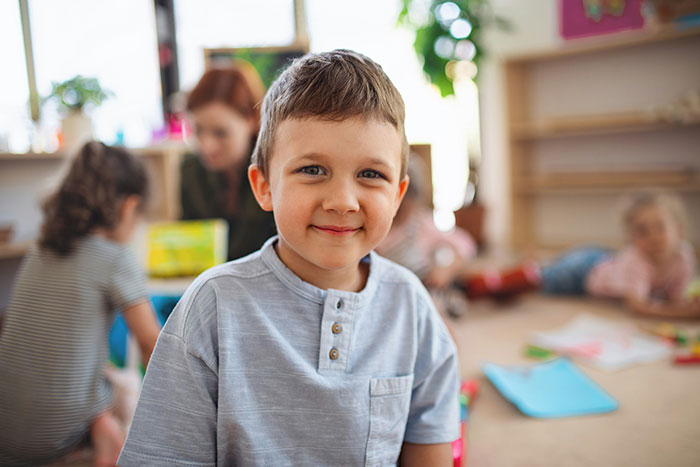 Young boy smiling in a classroom surrounded by kids, illustrating kids in school who earned the weirdo title.