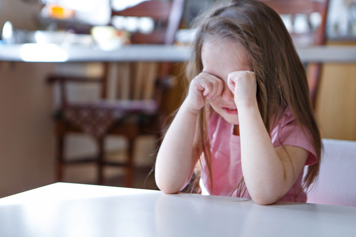 Young girl sitting at kitchen table, rubbing her eyes and upset, illustrating school suspending daughter over fear of snakes. Young girl sitting at kitchen table, rubbing her eyes and upset, illustrating school suspending daughter over fear of snakes.
