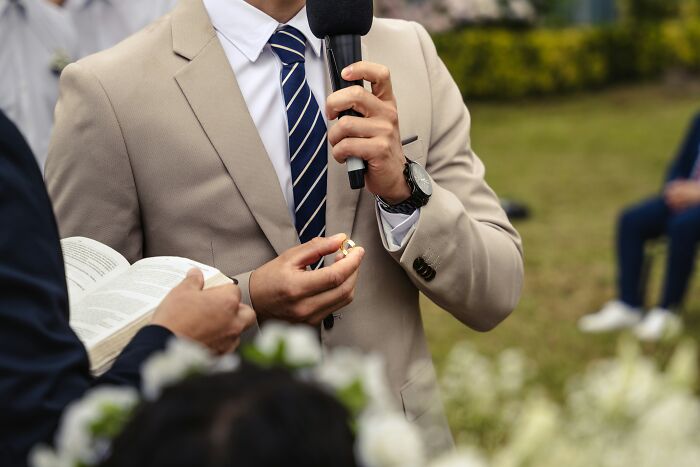 Man in beige suit holding wedding rings and microphone during a trashy, tacky, and distasteful wedding ceremony outdoors.