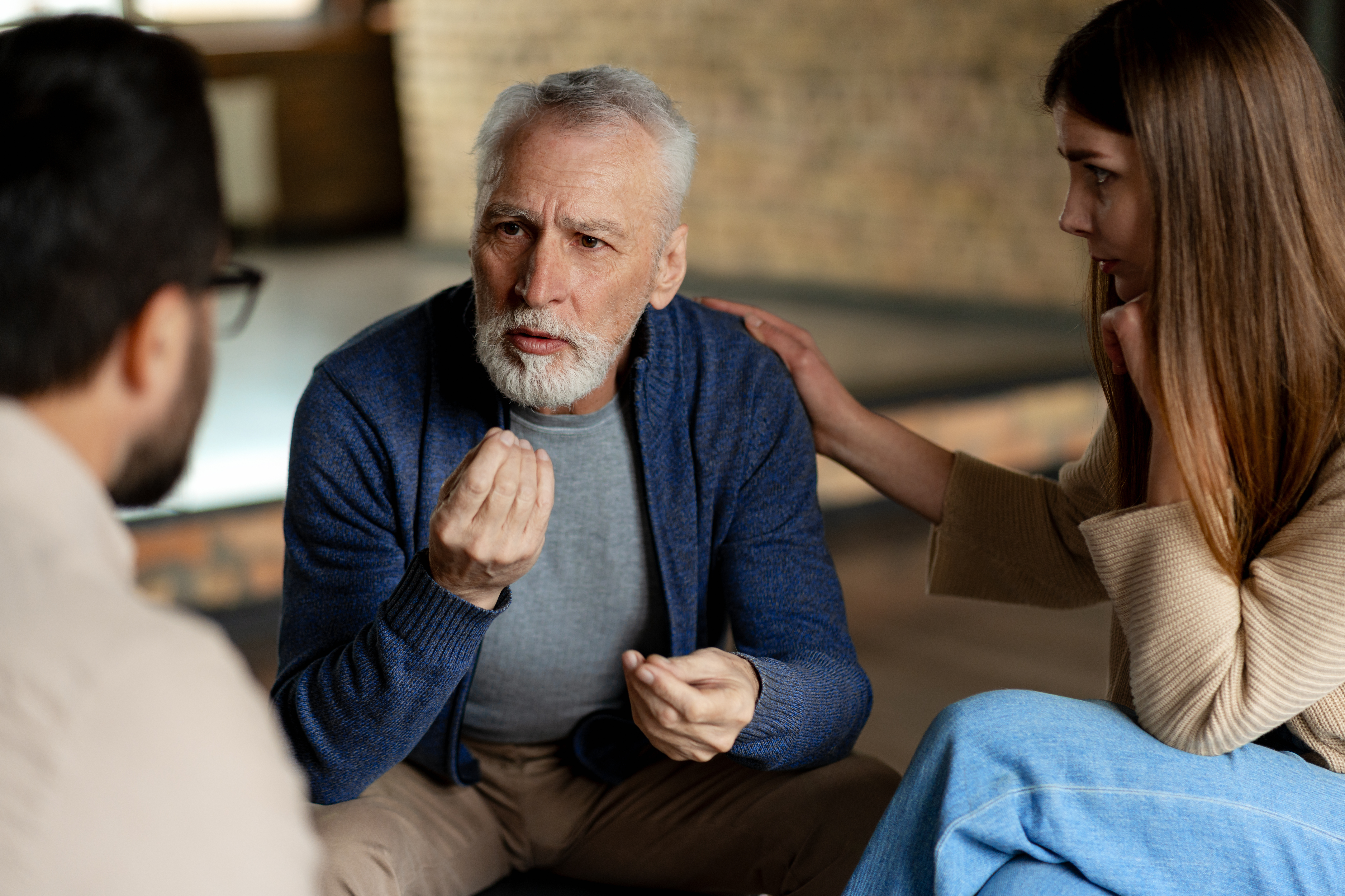 Older man confronting younger man about controlling behavior, while a woman supports the older man during a serious discussion.