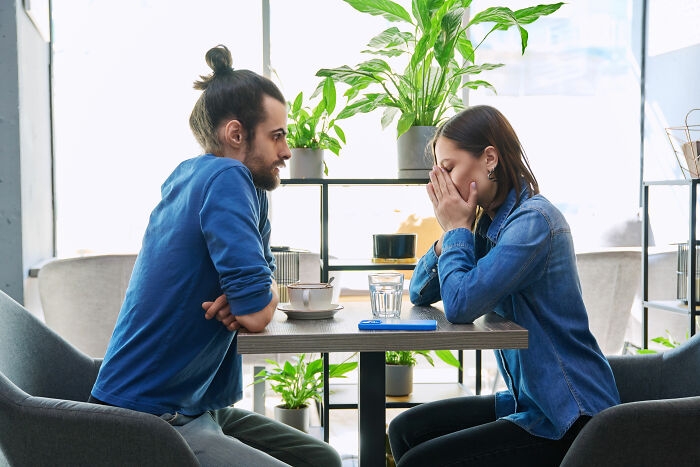 A man and a crying woman at a cafe. The man stares as the woman covers her face, possibly after finding out they were being cheated on.