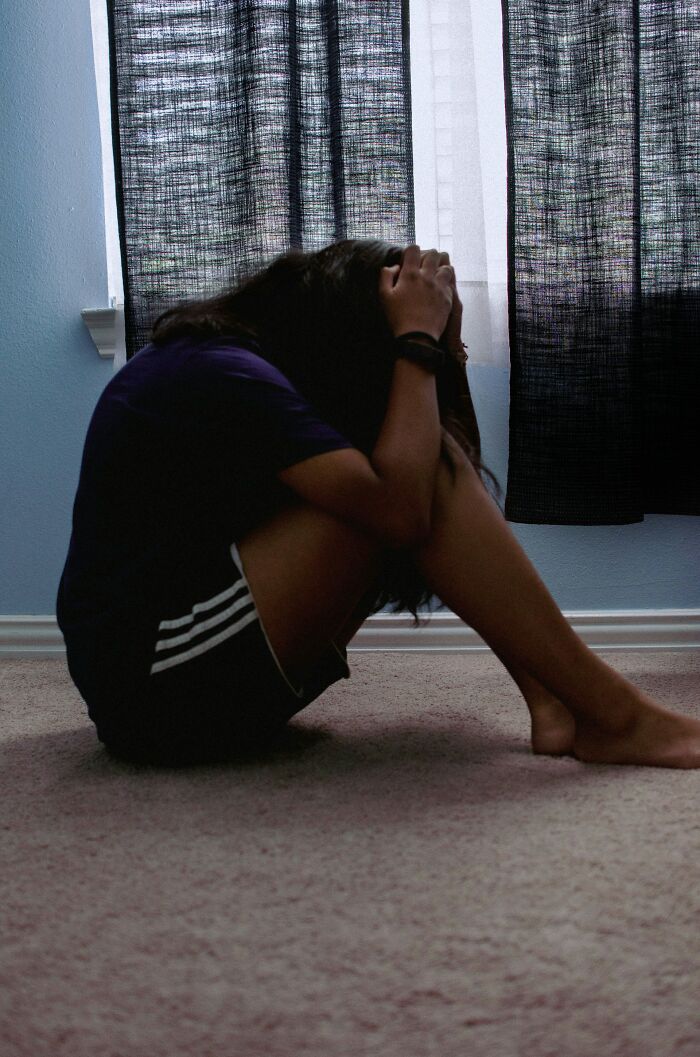 Person sitting on floor holding head in hands by window, reflecting on dark family secrets and emotional distress.