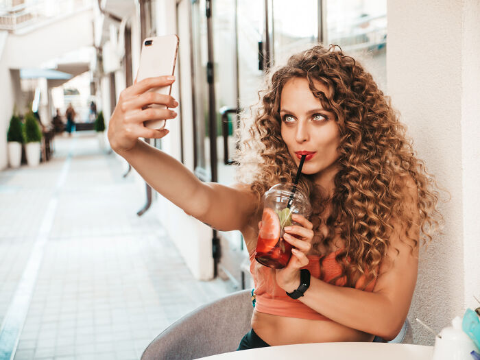 Young woman with curly hair taking a selfie while drinking a cocktail, related to infidelity stories getting exposed.