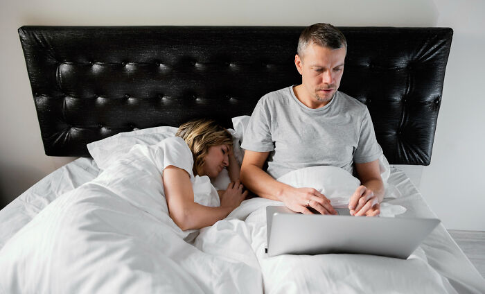 Man using laptop in bed while woman sleeps beside him, illustrating themes of infidelity getting exposed and accidental texts.
