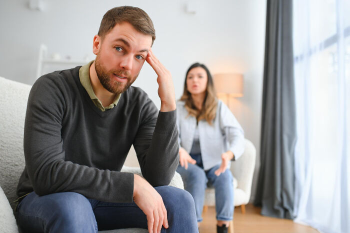 Man looking frustrated on couch while woman in background talks, illustrating infidelity getting exposed in relationships.