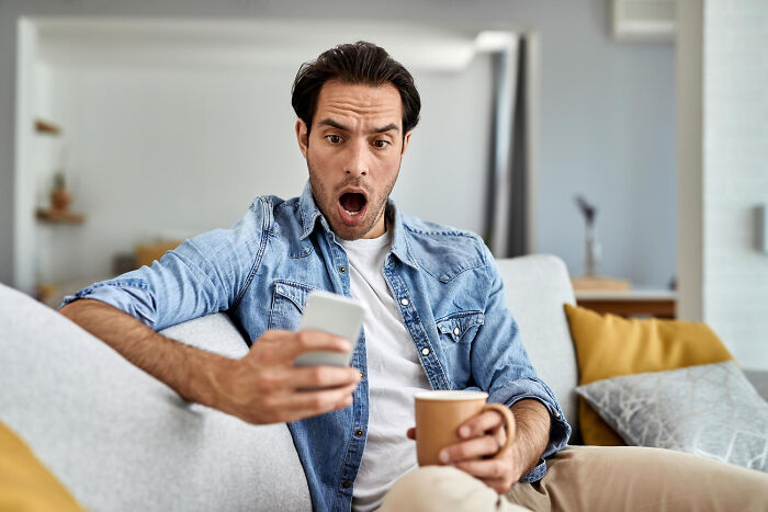Man sitting on couch holding phone and coffee with shocked expression representing infidelity getting exposed stories