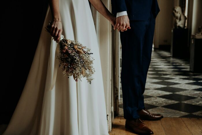 Bride in white dress holding dried flower bouquet and groom in navy suit holding hands at wedding venue before party