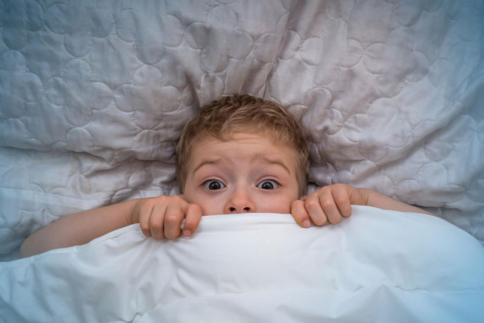 A scared 9yo boy peeks over a white blanket in bed, eyes wide with fear.