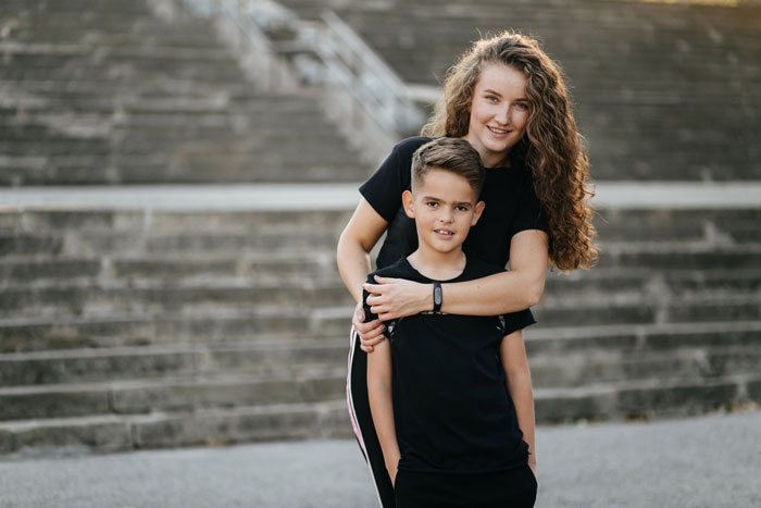 A smiling sister hugs her 9yo bro outdoors, both in black shirts, against a blurred concrete staircase.