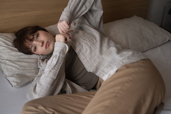A young person with short brown hair and a plaid shirt lies in bed, looking thoughtful. A scared brother might seek comfort.
