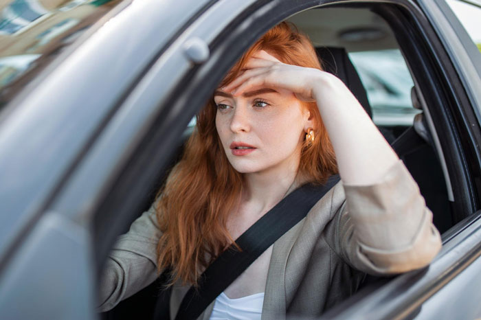 Young woman with red hair sitting in a car, looking concerned and shielding her eyes with her hand.