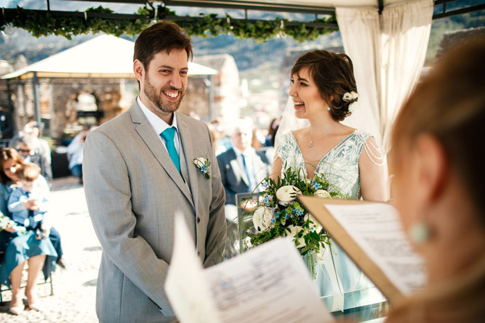 Bride and groom smiling during outdoor wedding ceremony as family members watch in the background on a sunny day