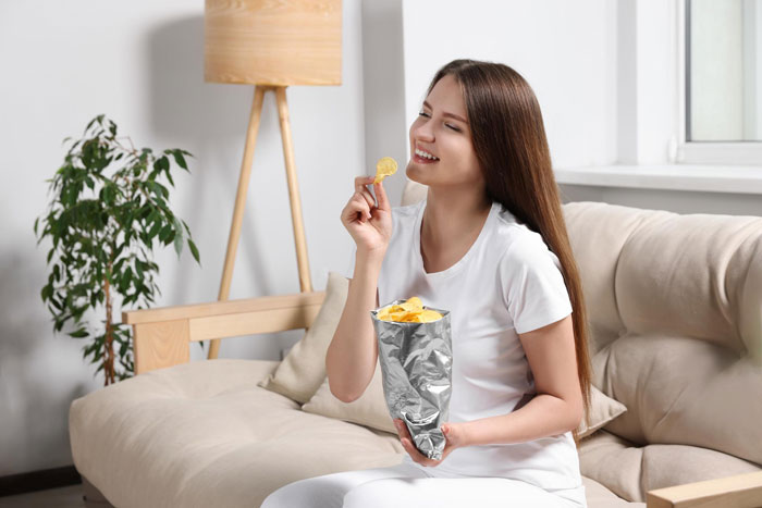 Young woman sitting on a couch eating chips, illustrating rude man snatching things without asking concept.