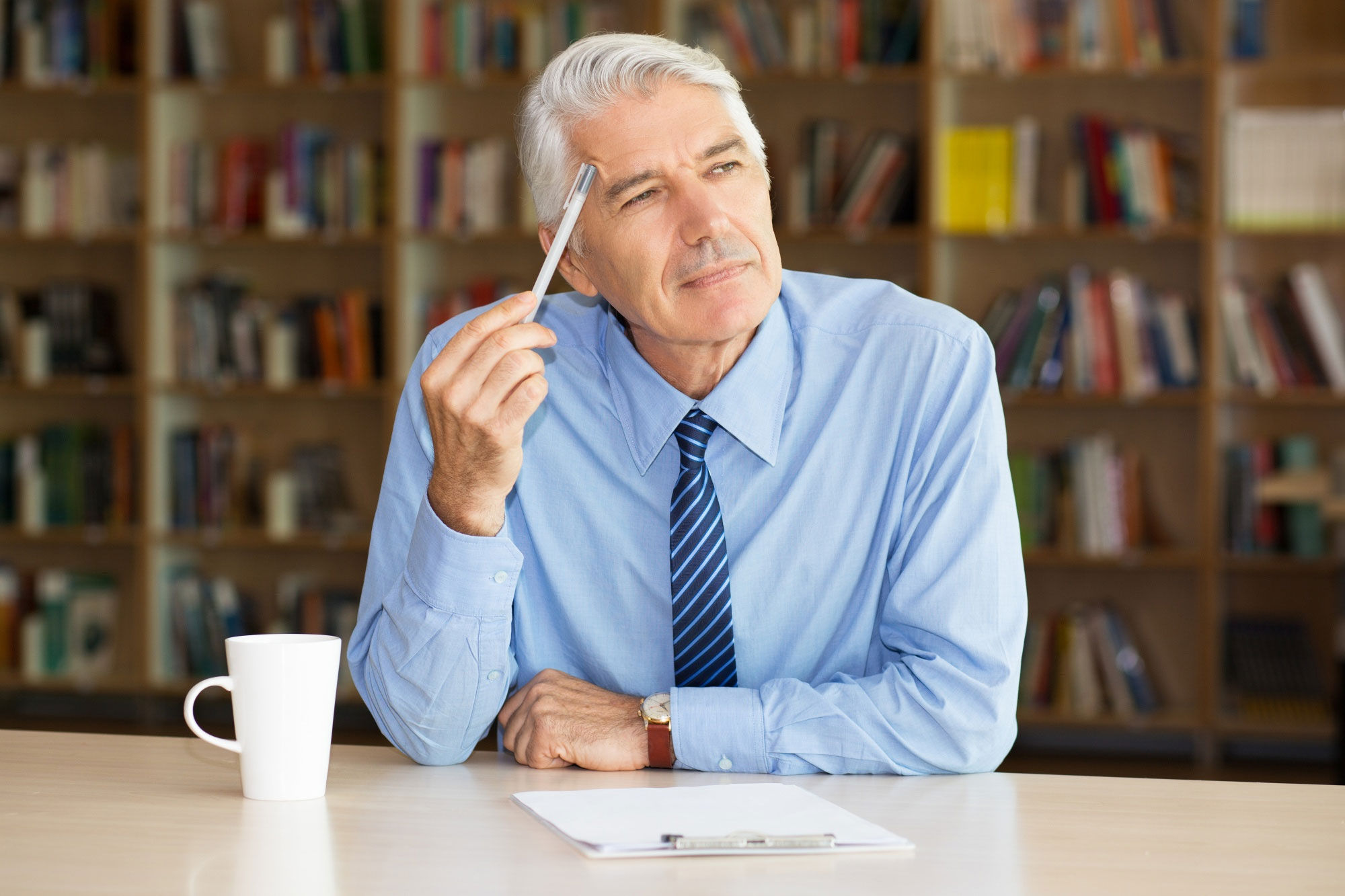 Older man in blue shirt and striped tie thinking while holding pen, with coffee cup and clipboard on desk in office library.