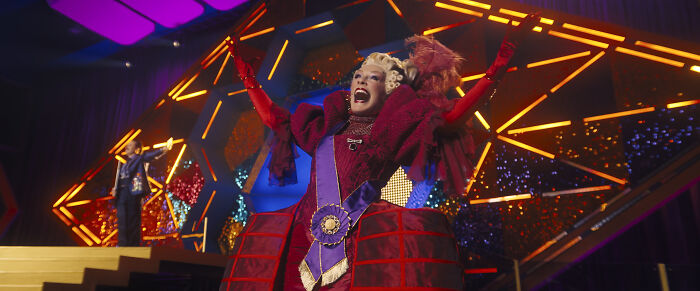 Actress in elaborate costume on stage reenacting iconic moment from The Hunger Games trailer with vivid lighting and audience.