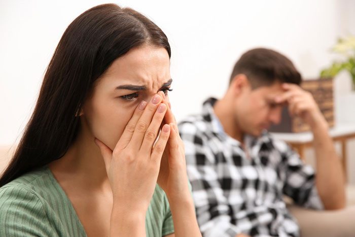 Young woman facing karma after cheating, covering mouth in distress while man in background looks frustrated and upset.