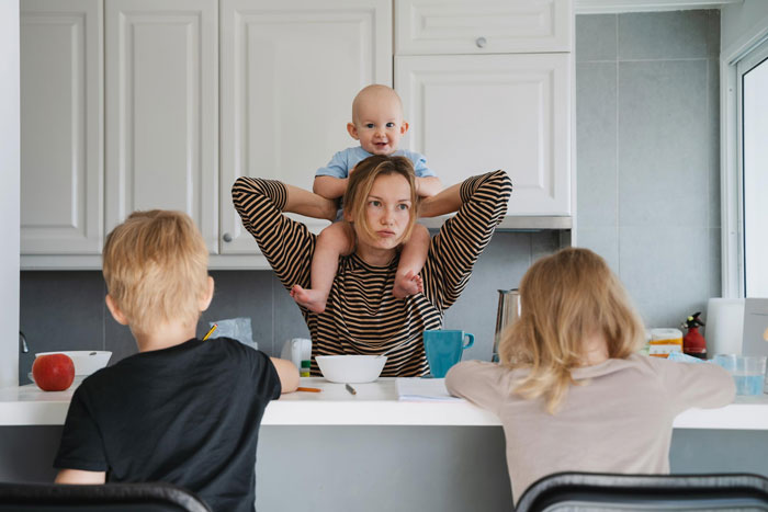 Woman facing karma with a baby on her shoulders and two children sitting at the kitchen counter in a modern home.