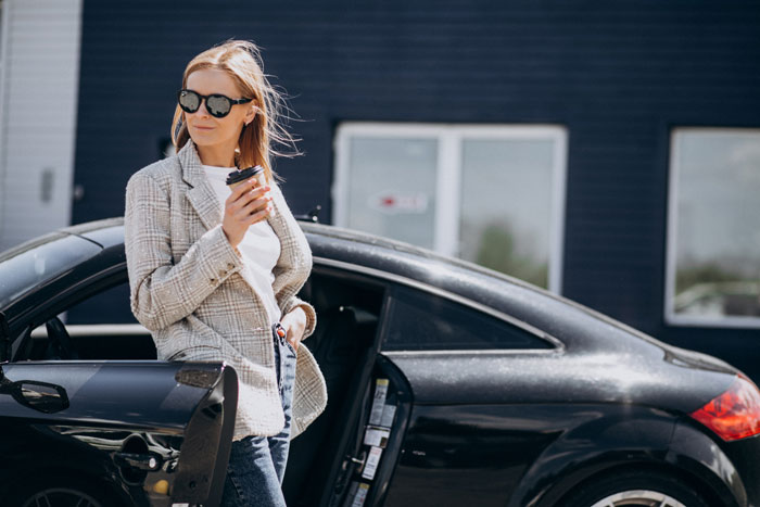 Woman standing by car door holding coffee, sunglasses on, showing confidence after trapping entitled guests in parking spot.