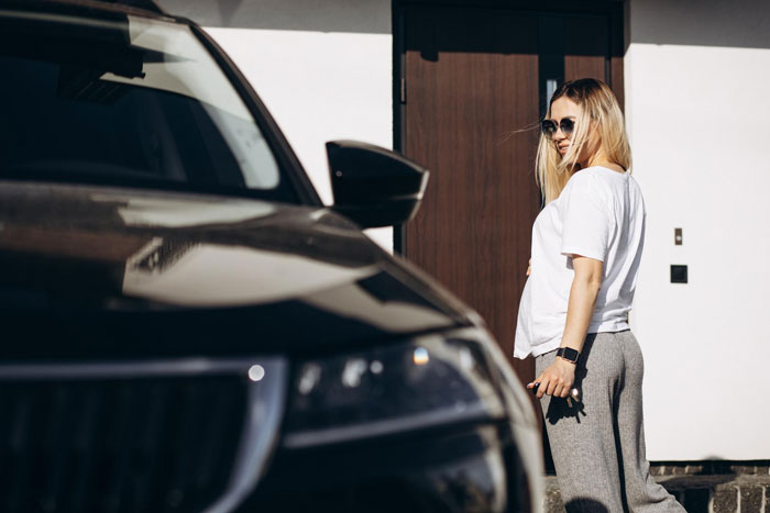 Woman standing near car parked in her reserved parking spot, wearing sunglasses and holding car keys confidently.