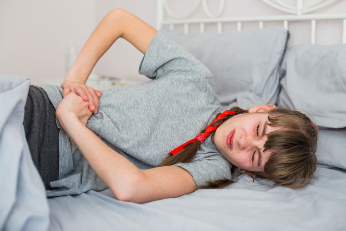 A young girl lying on her side in bed, clutching her stomach in pain. She is getting her period and looks distressed.