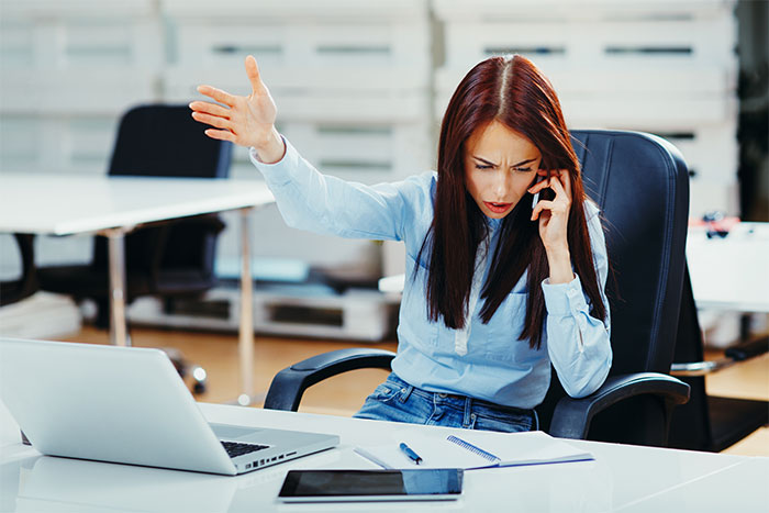 Woman on phone in office, looking frustrated, raising hand while sitting at desk with laptop and notebook, showing support.