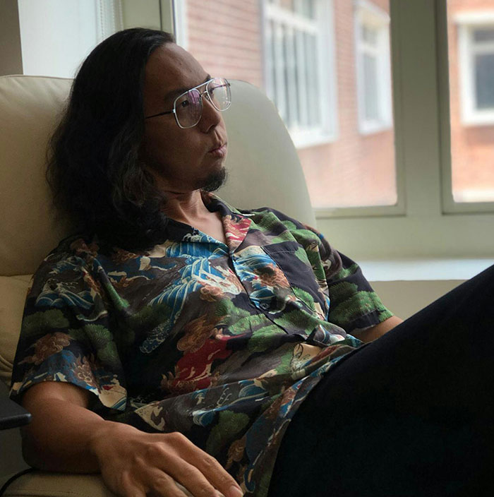 Man with long hair and glasses sitting thoughtfully in a chair, showing subtle and interesting displays of intelligence.