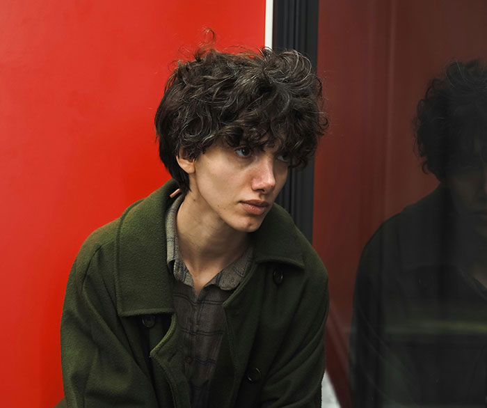 Young person with curly hair wearing green coat sitting thoughtfully next to red wall reflecting subtle displays of intelligence.
