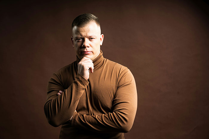 Man in a brown turtleneck posing thoughtfully against a dark brown background, showing subtle displays of intelligence.