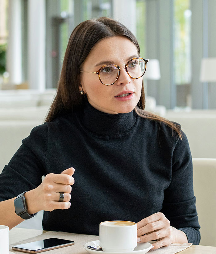 Woman wearing glasses and a black turtleneck showing subtle and interesting displays of intelligence during a coffee meeting.