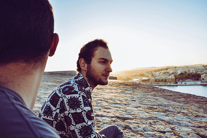 Two men sitting outdoors on a stone surface at sunset, engaged in a subtle and interesting display of intelligence.