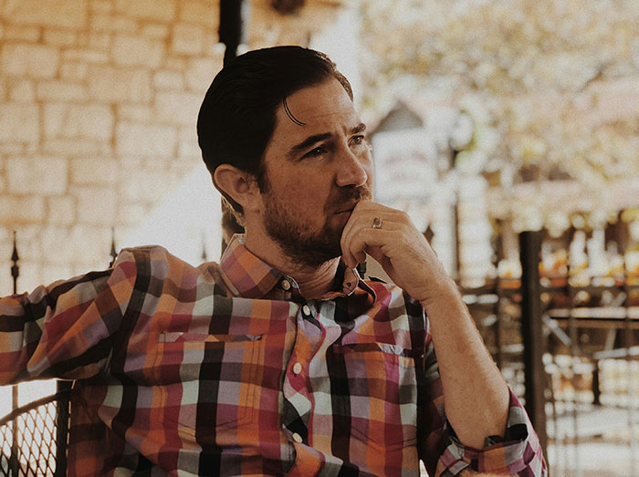 Man in a plaid shirt thoughtfully displaying subtle intelligence while seated in a relaxed outdoor setting.