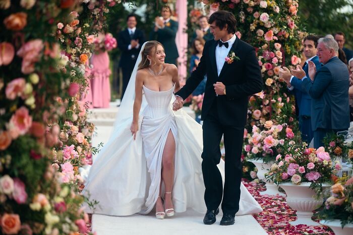 Sydney Sweeney, in character from Euphoria, smiles while walking hand-in-hand with a man down a flower-lined aisle.