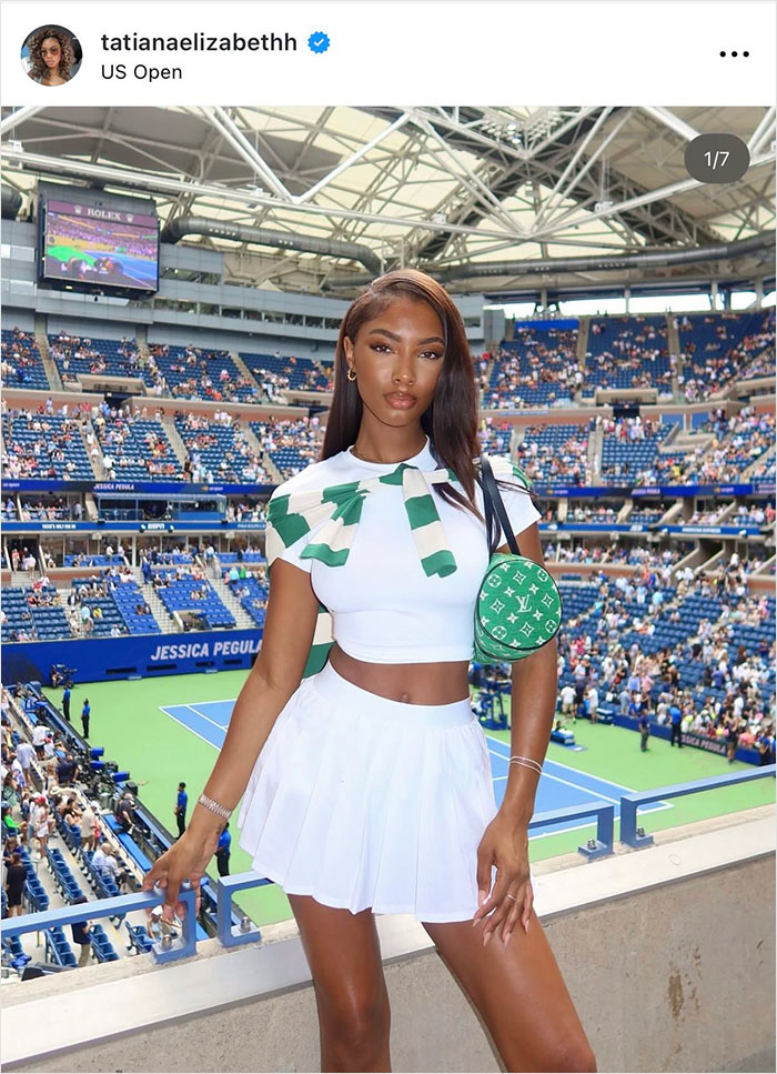 Glamorous influencer in white tennis outfit at US Open stadium with fans in the background noticing something off.