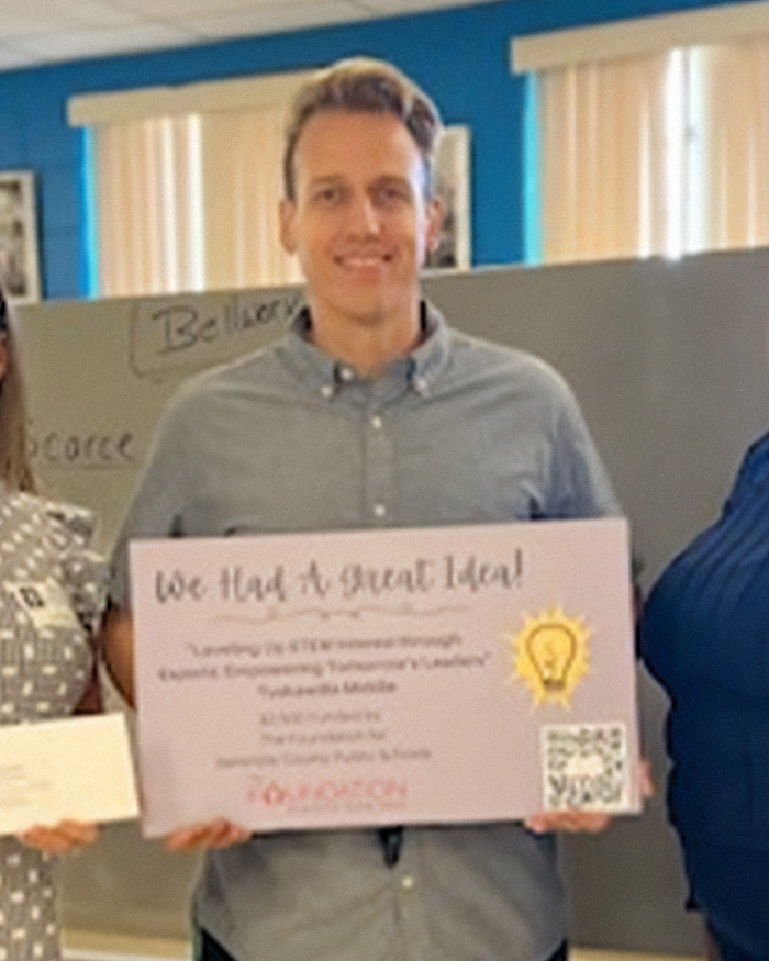 Homem segurando uma placa em uma sala de aula, relacionada a uma professora que supostamente está construindo um relacionamento com a mãe por um motivo sinistro.