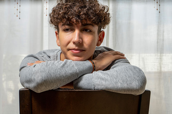 A young man with curly hair, in a gray sweater, resting his arms on a chair, looking thoughtful about a pregnant GF.