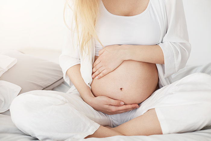 A pregnant woman in white clothing sits on a bed, cradling her baby bump with both hands.