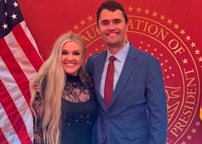 Erika Kirk sorrindo com um homem de terno em um evento formal com uma bandeira americana e um selo oficial ao fundo.