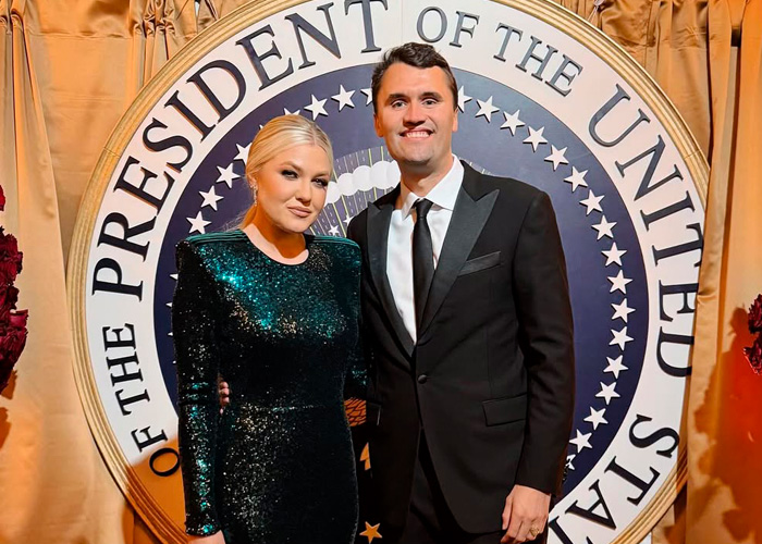Erika Kirk e um homem posando em traje formal em frente ao selo presidencial durante um evento de visita a uma escola secundária.