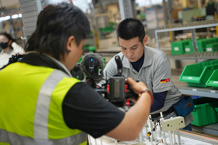 Worker going the extra mile being filmed at a manufacturing station while management watches in the background.
