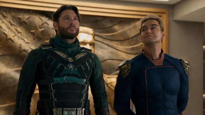 Dois personagens de The Boys em trajes de super-heróis. Menino Soldado (à esquerda) com barba e terno verde, Homelander (à direita) com cabelos loiros e terno azul. Eles olham para cima.