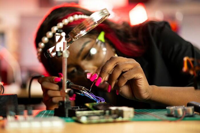A Black woman with pink nails, glasses, and a pearl headband solders a circuit board, exemplifying advice women give dads raising daughters.