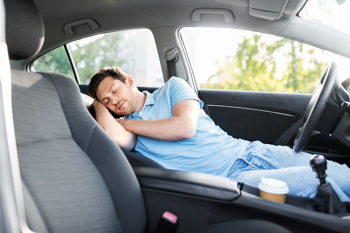 A man in a blue shirt and jeans sleeps inside a car with a coffee cup nearby, representing anonymous confessions & group therapy.