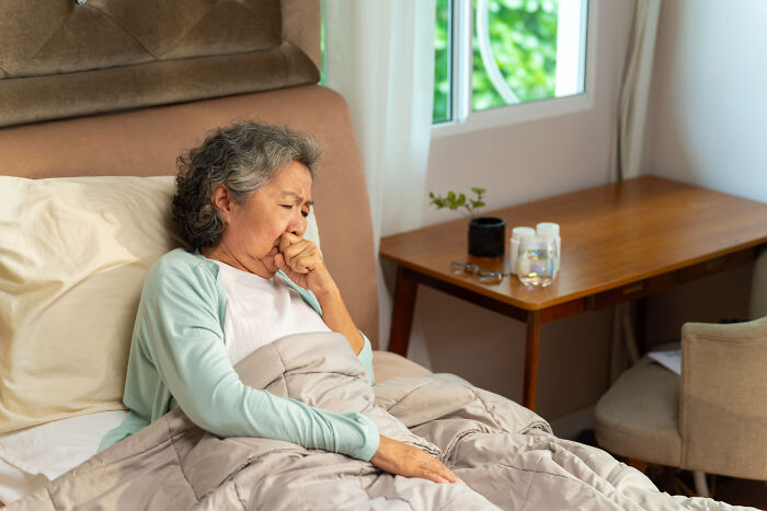 Elderly woman in bed coughing, appearing unwell in a cozy room, illustrating the darkest turn of being late.