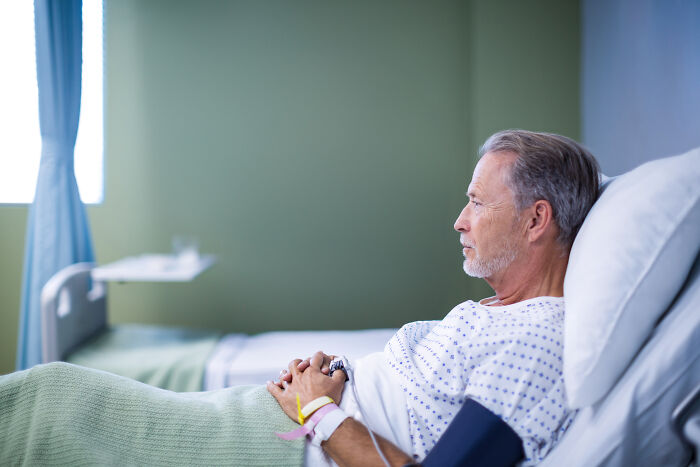 Elderly man in hospital bed wearing patient gown, appearing contemplative after a late arrival causing serious consequences.