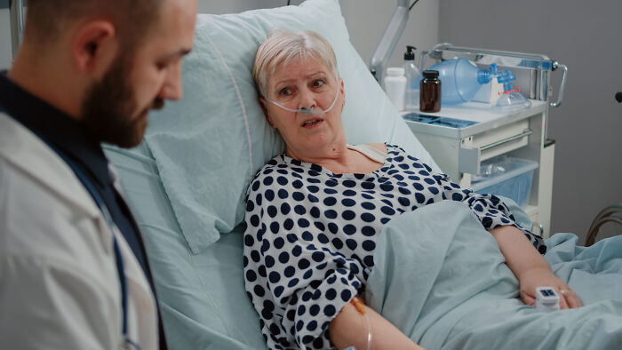 Elderly woman in hospital bed with oxygen tube talking to concerned doctor about late arrival consequences.